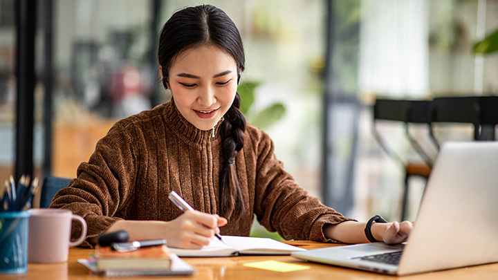 A woman smiling as she takes notes in a notebook while working on a laptop at a desk.