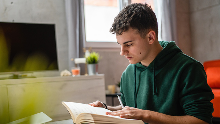 A young man in a green hoodie sitting indoors, focused on reading a large open book and holding a pen.