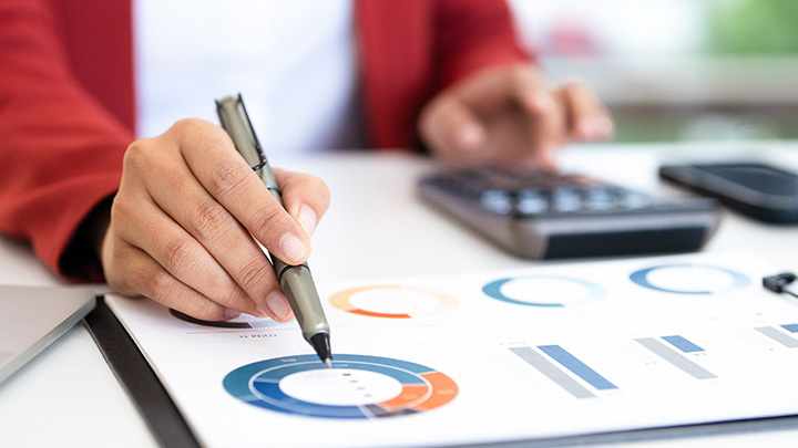 A person in a red jacket annotates printed financial charts and graphs at a desk, with a calculator nearby.