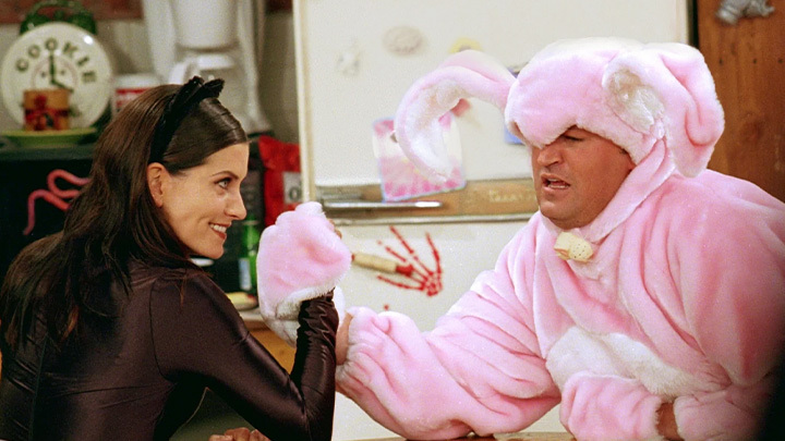 A shot taken from the tv show, Friends, of Chandler Bing dressed in a pink rabbit costume who appears to be struggling while having an arm wrestle with character, Monica Geller, dressed in a black cat woman costume.