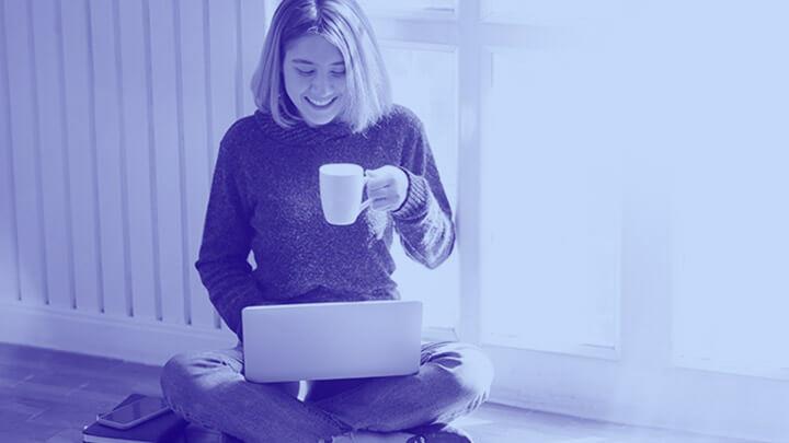 Student sat on floor with laptop and mug in hand, smiling