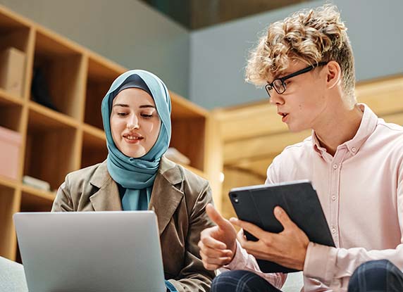 A woman using a laptop talking to a man holding a tablet computer.