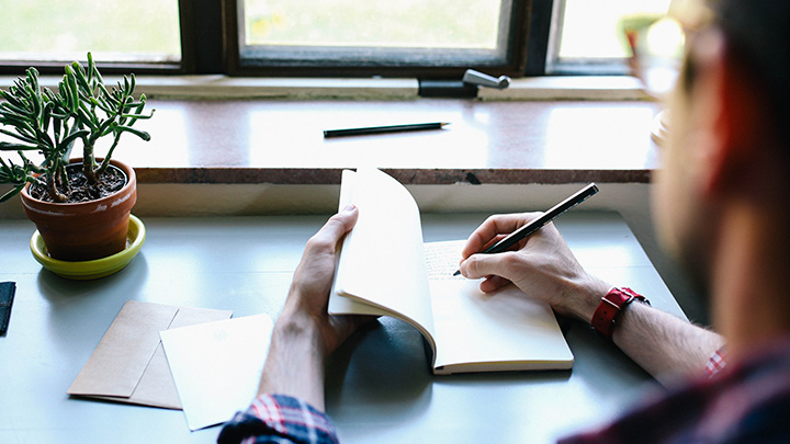 A person sitting at a desk in the window writing in a notebook.