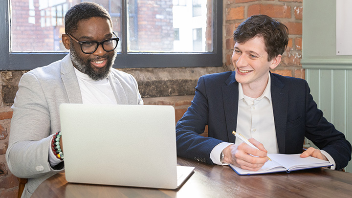 Two men looking at a laptop screen. One man is holding a pen and a notebook.