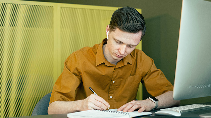 A man writing in a notebook in front of a computer.