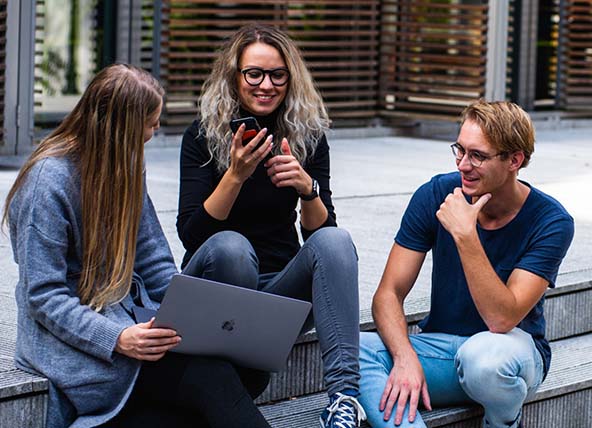 A woman looking at her phone talking to a woman using a laptop, and a man, who are all sat down on some steps