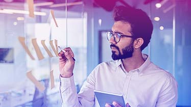A man wearing glasses looking at a post it-notes on a glass wall.
