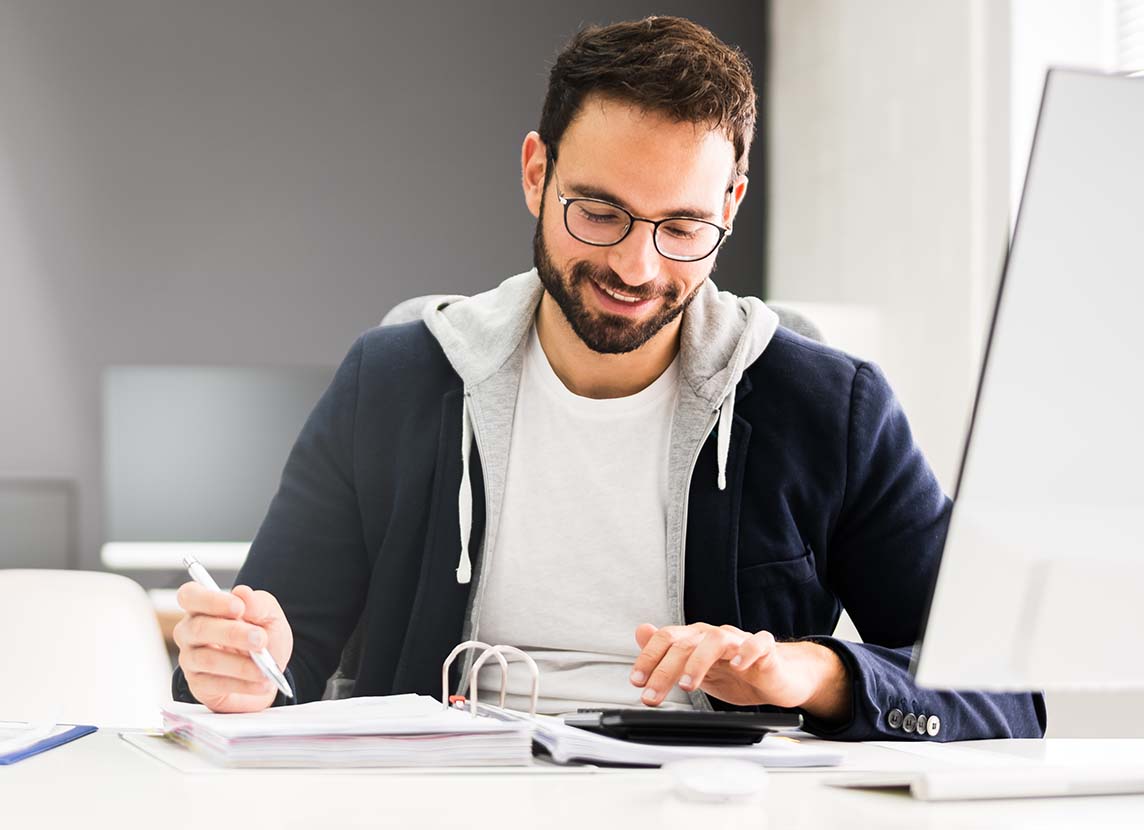 Man wearing glasses and a blazer sits at a desk, writing in a binder and using a calculator, with a computer monitor in the foreground.