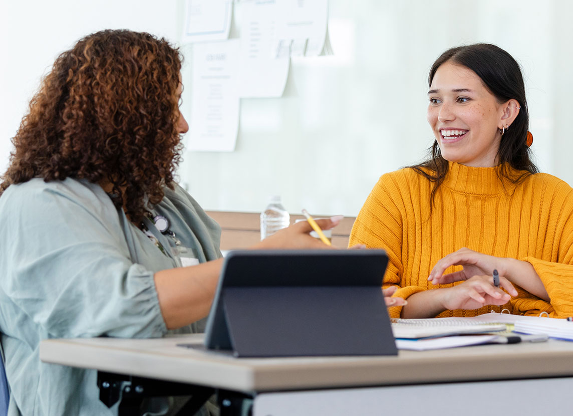 Two women sit at a table engaged in a friendly conversation. One is wearing a yellow sweater, smiling, as they discuss papers and a tablet.