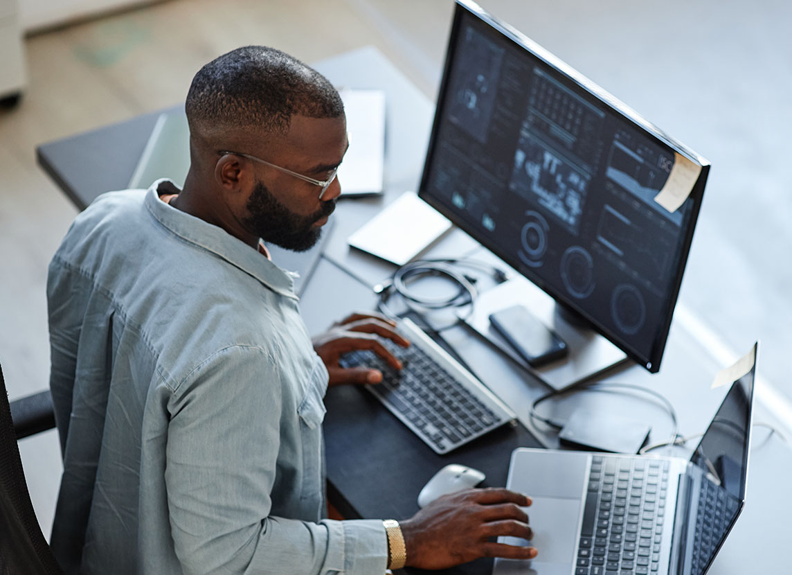 Man sitting at a desk working on a computer with multiple screens displaying data and graphs. The setting is modern and focused, conveying concentration.