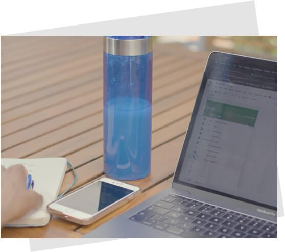 A hand holding a pen on a notebook next to a mobile phone, a water bottle and a laptop on a table