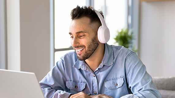 A man wearing headphones, smiling and working on a laptop.
