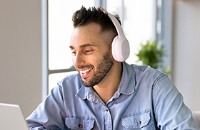 A man wearing headphones, smiling and working on a laptop.