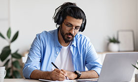 Man wearing headphones using a laptop and making notes
