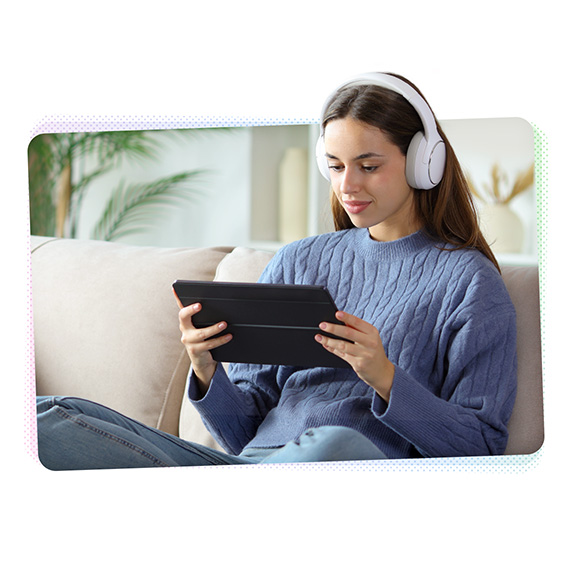 A woman sitting cross-legged on a sofa wearing headphones and looking at a tablet computer